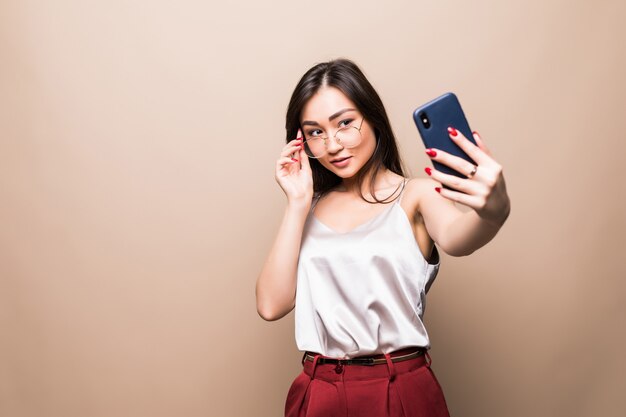 La muchacha bastante asiática toma el selfie con su teléfono inteligente aislado en la pared beige.