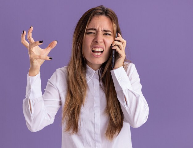 La muchacha bastante caucásica joven disgustada habla por teléfono y se coloca con la mano levantada en púrpura
