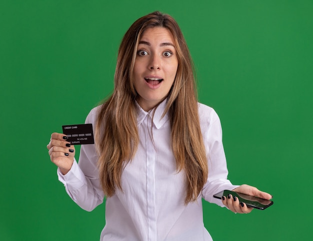 La muchacha bastante caucásica joven sorprendida sostiene la tarjeta de crédito y el teléfono aislados en la pared verde con el espacio de la copia