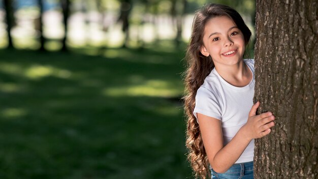 Muchacha bastante linda que mira a escondidas de tronco de árbol en el parque