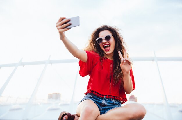 La muchacha bonita alegre con el pelo rizado toma un selfie en el teléfono móvil