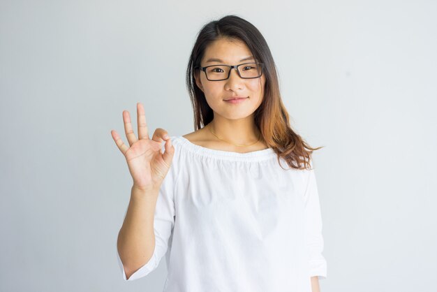 Muchacha bonita asiática del contenido con el pelo destacado que hace la muestra aceptable como símbolo de la aprobación.