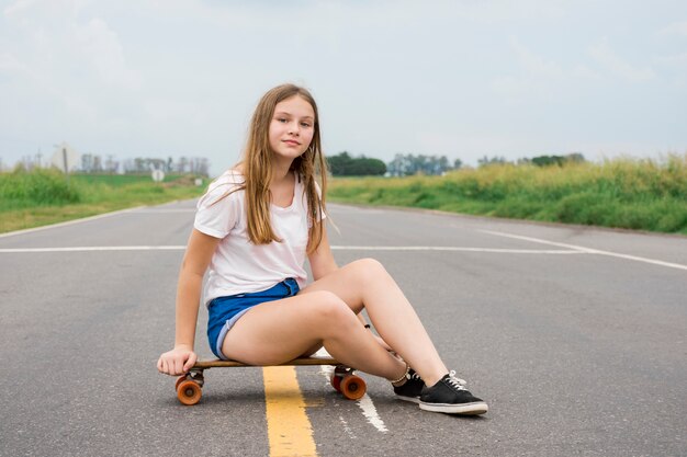 Muchacha bonita atractiva moderna que se sienta en el monopatín en el camino vacío