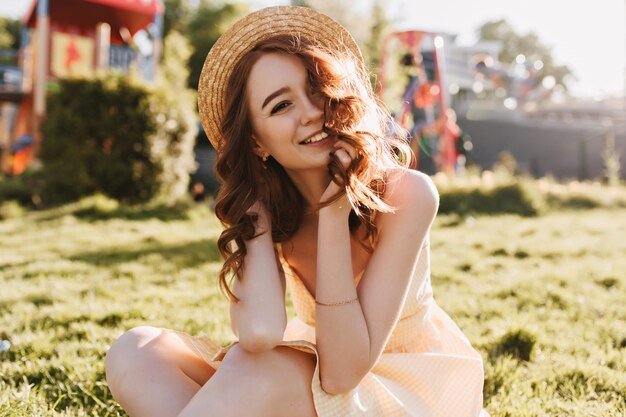 Muchacha bonita del jengibre en vestido amarillo que presenta en la hierba. Riendo a dama romántica con sombrero sentado en el césped verde.