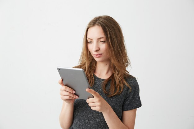 Muchacha bonita joven que sonríe mirando la tableta que navega el web de la navegación por Internet.