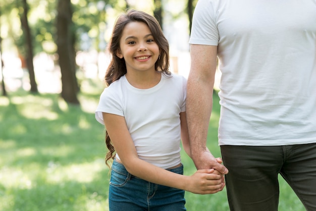 Muchacha bonita que mira la cámara que lleva a cabo la mano de su padre en parque