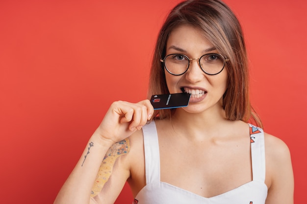 Muchacha bonita que sostiene con la mano una tarjeta de crédito plástica entre sus dientes en la pared roja.