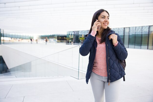 Muchacha bonita sonriente con la taleguilla usando el teléfono móvil al aire libre