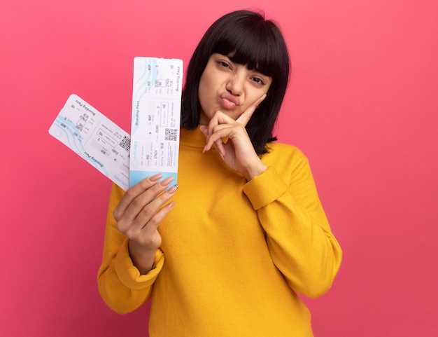 La muchacha caucásica morena joven descontenta pone la mano en la barbilla y sostiene los billetes de avión en rosa