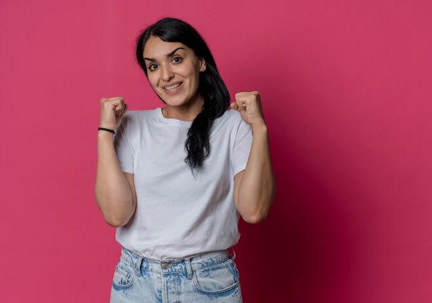 La muchacha caucásica morena joven emocionada mantiene los puños mirando aislados en la pared rosada