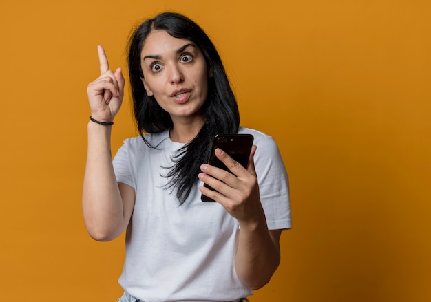 La muchacha caucásica morena joven sorprendida apunta hacia arriba sosteniendo el teléfono aislado en la pared naranja