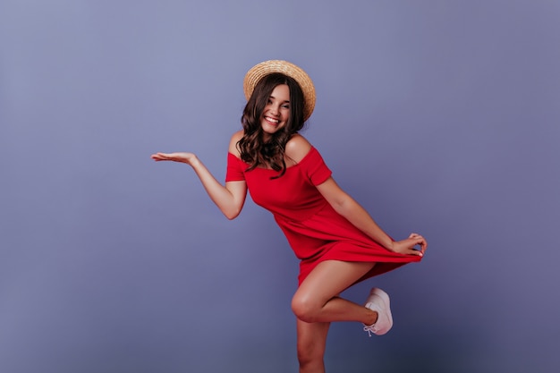 Muchacha caucásica en sombrero de paja que presenta en la pared púrpura. Sonriente mujer encantadora en vestido rojo bailando.