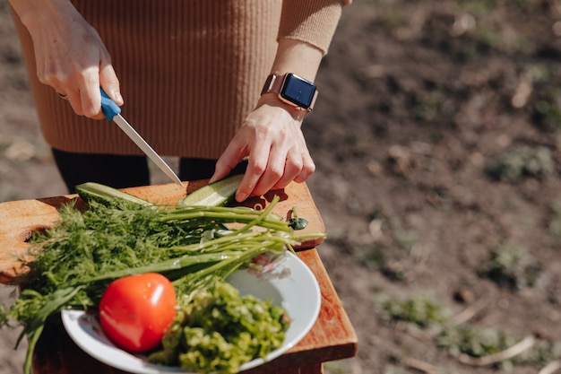 Foto gratuita la muchacha corta verduras en el tablero y prepara una ensalada en la naturaleza. día soleado y cocina. vista de primer plano.