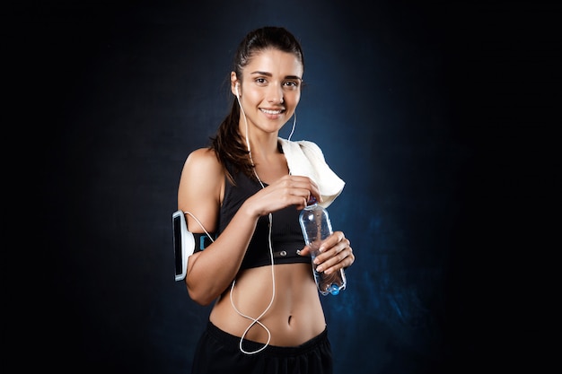 Muchacha deportiva hermosa joven que sostiene el agua sobre la pared oscura.