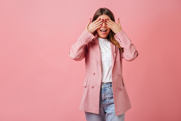 Muchacha divertida en la chaqueta casual que cubre los ojos. Foto de estudio de dama de moda aislada sobre fondo rosa.