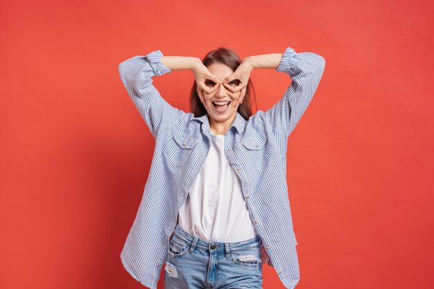 Muchacha divertida, linda que se divierte aislada en una pared roja mientras que hace las manos de los prismáticos.