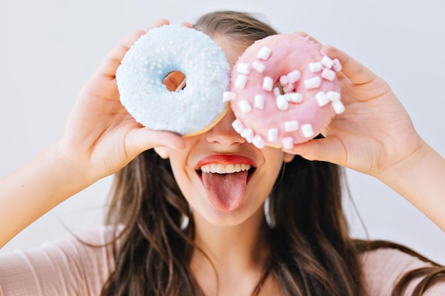 Muchacha divertida del retrato del primer con el pelo largo que se divierte con las donas coloridas contra sus ojos. Mujer joven atractiva con labios rojos mostrando la lengua. Expresiones, concepto de dieta, colores brillantes