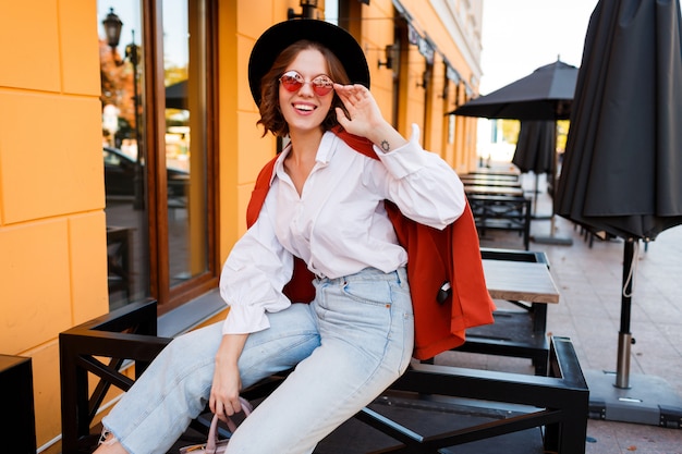 Muchacha europea sonriente en lindas gafas de sol naranjas, chaqueta y sombrero negro sentado al aire libre.