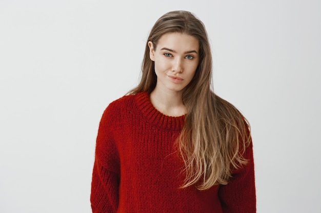 La muchacha expresa desdén al rival. Retrato de mujer hermosa no impresionada disgustada en suéter rojo suelto de moda, sonriendo con desprecio, dudando si podría confiar en la persona