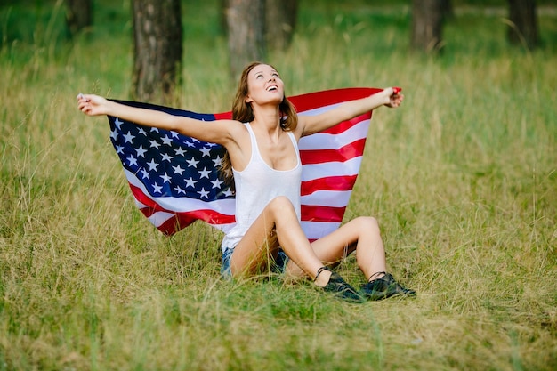 La muchacha feliz se está sentando en la hierba con la bandera americana