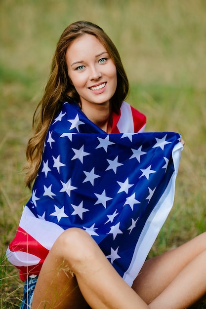 La muchacha feliz se está sentando en la hierba con la bandera americana