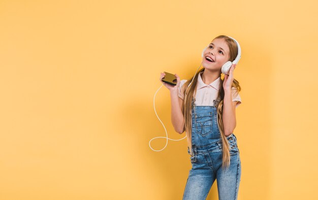 Muchacha feliz que disfruta de la música en el auricular que sostiene el teléfono móvil en la mano que se opone a fondo amarillo