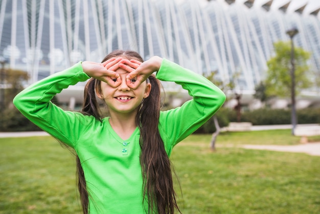 Foto gratuita muchacha feliz que forma las gafas con su dedo que se coloca en parque