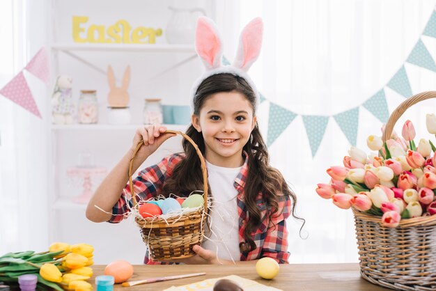 Muchacha feliz que lleva los oídos del conejito que sostienen la cesta de los huevos de Pascua coloridos