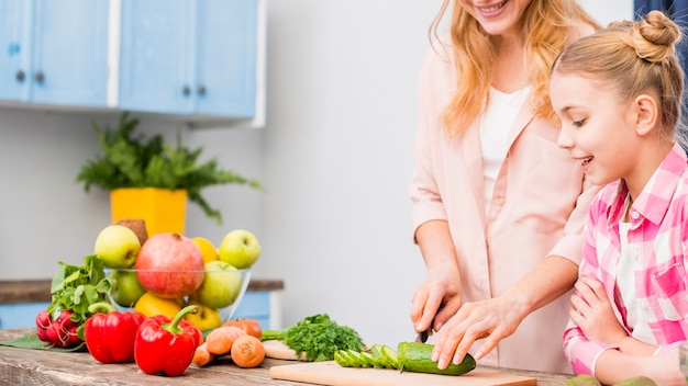 Muchacha feliz que mira a su madre que corta la rebanada del pepino con el cuchillo en la tabla