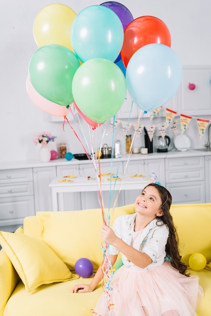 Muchacha feliz que se sienta en el sofá que sostiene los globos coloridos