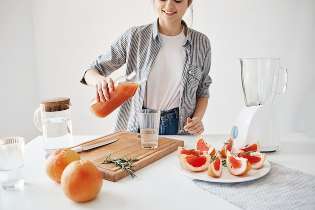 Muchacha hermosa feliz que vierte el smoothie de desintoxicación del pomelo en el vidrio que sonríe sobre la pared blanca. Dieta saludable nutrición.