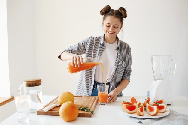 Muchacha hermosa feliz que vierte el smoothie de desintoxicación del pomelo en el vidrio que sonríe sobre la pared blanca. Dieta saludable nutrición.