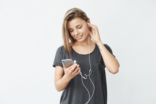 Muchacha hermosa joven en auriculares que sonríe mirando la pantalla del teléfono.