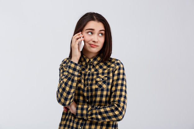 La muchacha hermosa joven irónica se vistió en camisa de tela escocesa que hablaba en el teléfono, mirando el lado sobre la pared blanca.