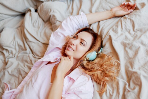 La muchacha hermosa joven del jengibre en auriculares que se estira en cama después de despertar descansó completamente.