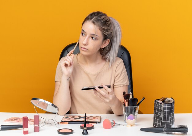 La muchacha hermosa joven del lado que mira impresionado se sienta a la mesa con las herramientas de maquillaje que sostienen la paleta de la sombra de ojos con el cepillo de maquillaje