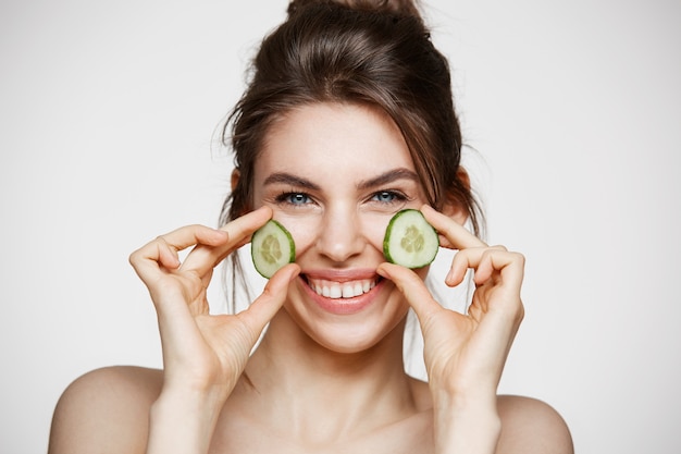 Muchacha hermosa joven con la piel limpia perfecta que sonríe mirando la cámara que sostiene rebanadas del pepino sobre el fondo blanco. Cosmetología de belleza y spa.