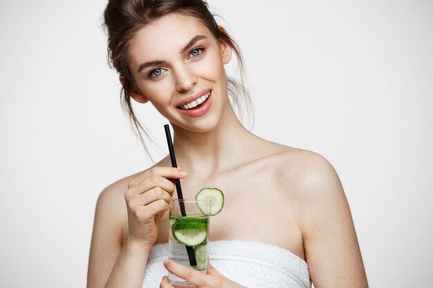 Muchacha hermosa joven con la piel limpia perfecta que sonríe mirando la cámara que sostiene el vidrio de agua con las rebanadas del pepino sobre el fondo blanco. Nutrición saludable.