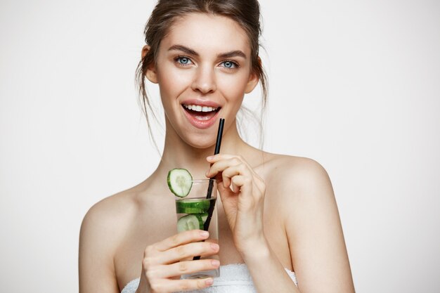 Muchacha hermosa joven con la piel limpia perfecta que sonríe mirando la cámara que sostiene el vidrio de agua con las rebanadas del pepino sobre el fondo blanco. Nutrición saludable.