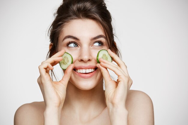 Muchacha hermosa joven con la piel limpia perfecta que sonríe sosteniendo las rebanadas del pepino sobre el fondo blanco. Cosmetología de belleza y spa.