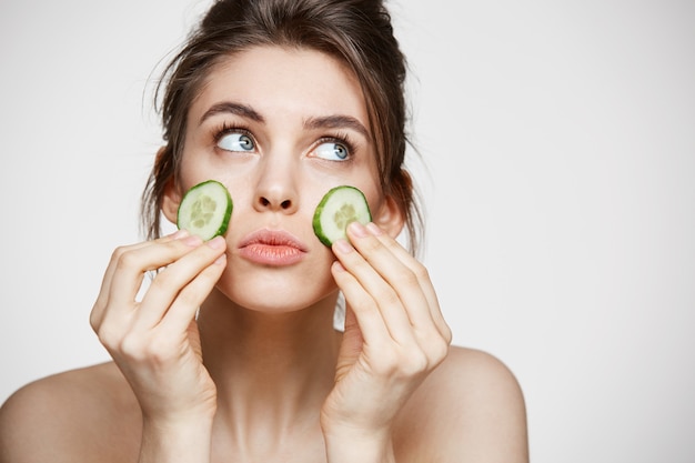 Muchacha hermosa joven con la piel limpia perfecta que sostiene rebanadas del pepino sobre el fondo blanco. Cosmetología de belleza y spa.