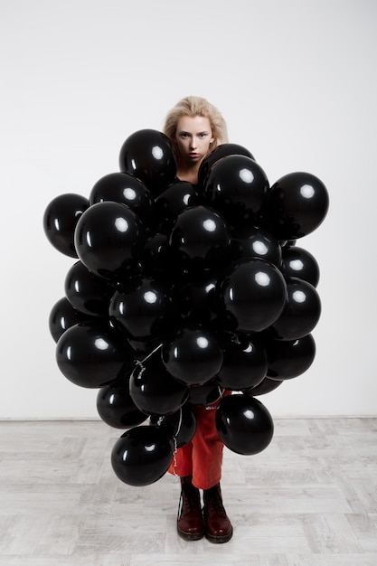 Muchacha hermosa joven que se coloca en globos negros sobre la pared blanca.