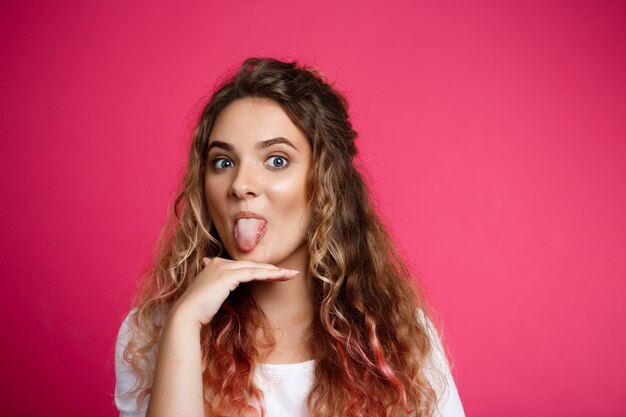 Muchacha hermosa joven que muestra la lengua sobre la pared rosada.