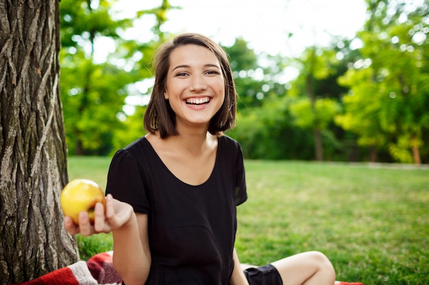 Muchacha hermosa joven que sonríe, sosteniendo la manzana en comida campestre en parque.