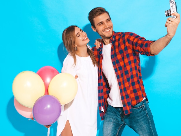 Foto gratuita muchacha hermosa sonriente y su novio hermoso que sostiene el manojo de globos coloridos. feliz pareja tomando foto selfie de sí mismos en la cámara retro. feliz cumpleaños