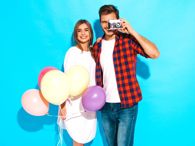 Muchacha hermosa sonriente y su novio hermoso que sostiene el manojo de globos coloridos. Feliz pareja tomando fotos de sí mismos en la cámara retro. Feliz cumpleaños