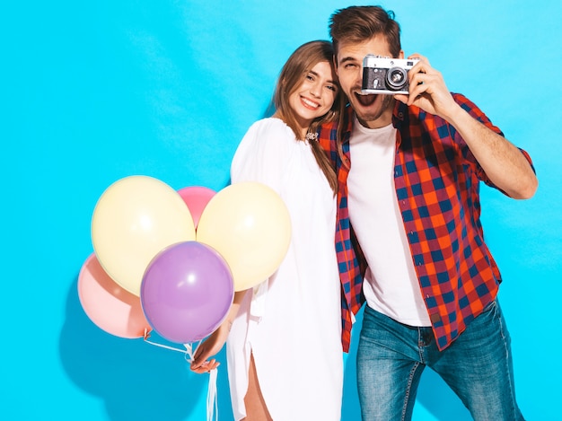 Muchacha hermosa sonriente y su novio hermoso que sostiene el manojo de globos coloridos. Feliz pareja tomando fotos de sí mismos en la cámara retro. Feliz cumpleaños