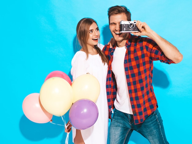 Muchacha hermosa sonriente y su novio hermoso que sostiene el manojo de globos coloridos. Feliz pareja tomando fotos de sí mismos en la cámara retro. Feliz cumpleaños