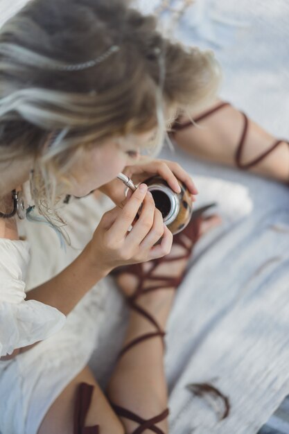 Muchacha india hermosa del hippie con el pelo rubio largo en el té de consumición del compañero del tejado.