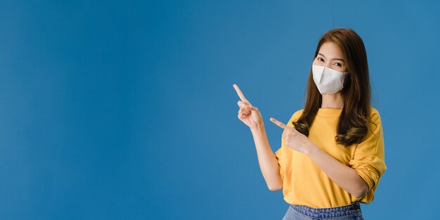 La muchacha joven de Asia usa una mascarilla médica que muestra algo en el espacio en blanco con vestida con un paño informal y mira a la cámara. Distanciamiento social, cuarentena por coronavirus. Bandera panorámica de fondo azul.